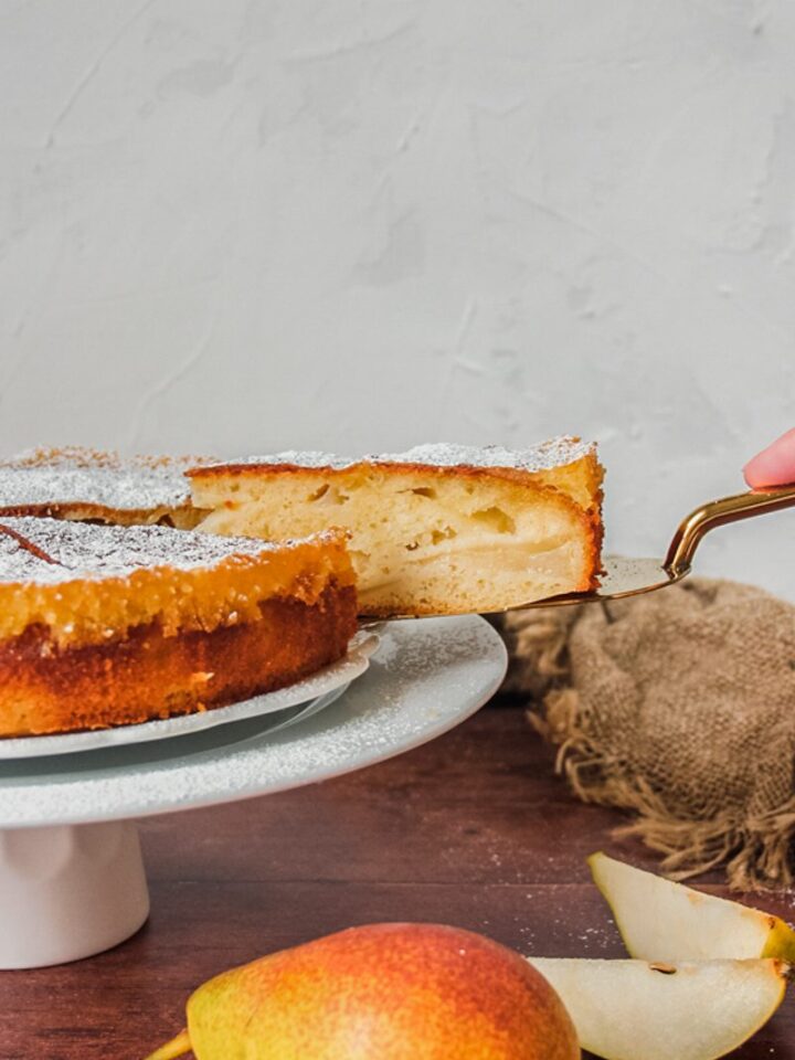 A slice of French pear cake is lifted from the cake plate sitting on a cake stand on a wooden background with a brown cloth in the background and pear in the foreground.