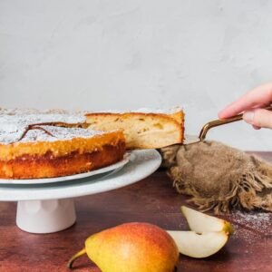 A slice of French pear cake is lifted from the cake plate sitting on a cake stand on a wooden background with a brown cloth in the background and pear in the foreground.