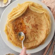 Jam is spread over a crepe like German pancake stack on a white ceramic plate on a gray surface.