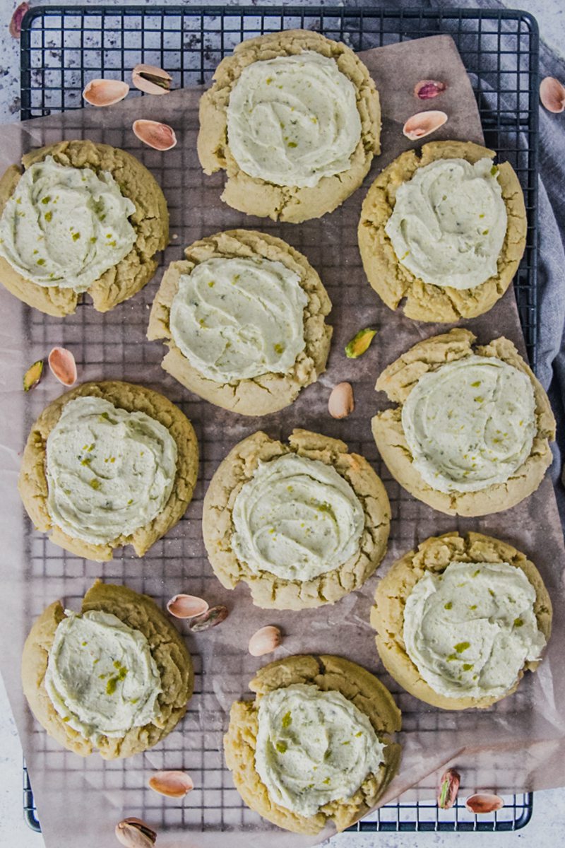 Pistachio sugar cookies with a pistachio buttercream sit on a parchment paper lined cooling rack above a light gray surface.