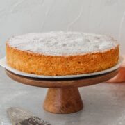 A plain sponge cake sits on a cake stand above a gray surface with a sprinkle of confectioner's sugar on top.
