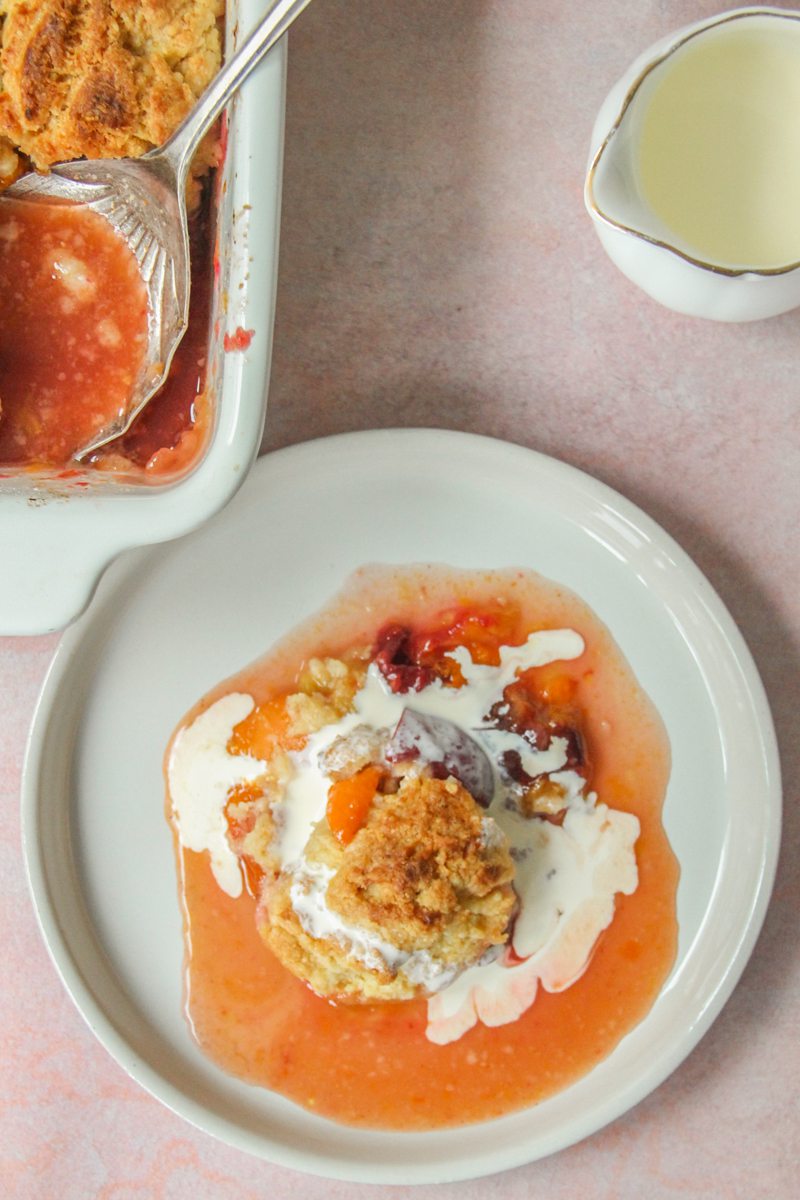 An individual serving of plum and apricot cobbler sits on a ceramic plate with a lip on a light pink surface.