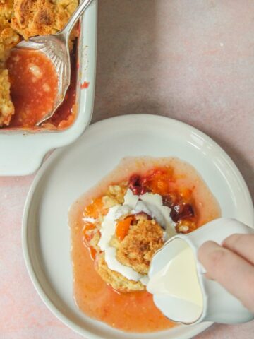 Cream is poured over the top of an individual serving of plum and apricot cobbler on a ceramic plate on a pink surface.