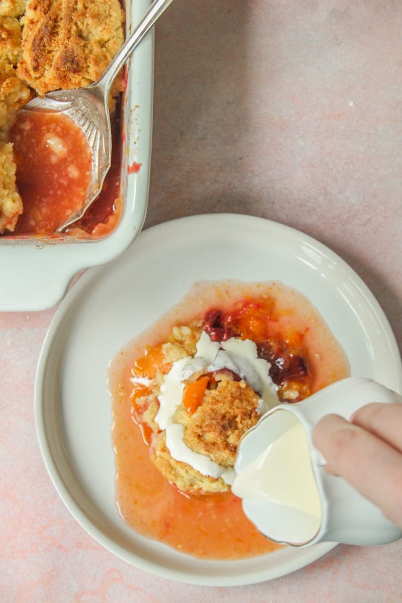Cream is poured over the top of an individual serving of plum and apricot cobbler on a ceramic plate on a pink surface.