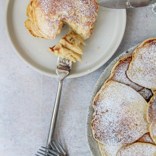 Polish Apple Pancakes - Racuchy z Jablkami sit on an individual plate with a portion on a fork resting on the edge of the plate and the remaining pancakes beside on a larger plate on a gray surface.