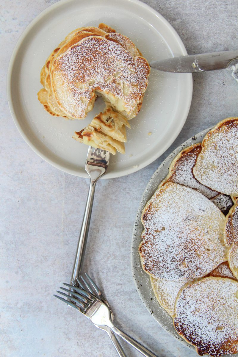 Polish Apple Pancakes - Racuchy z Jablkami sit on an individual plate with a portion on a fork resting on the edge of the plate and the remaining pancakes beside on a larger plate on a gray surface.