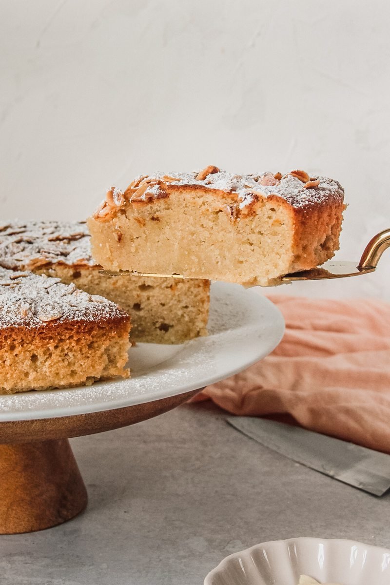 A slice of Portuguese Almond Cake sits on a cake server with the remaining cake on a cake stand on a light gray surface.