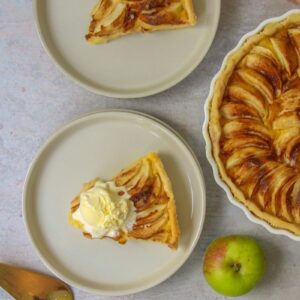 A slice of Portuguese Apple Tart sits on a rimmed ceramic plate with a scoop of vanilla ice cream on top. A second slice sits nearby without alongside the remaining tart, server and apple on a gray surface.