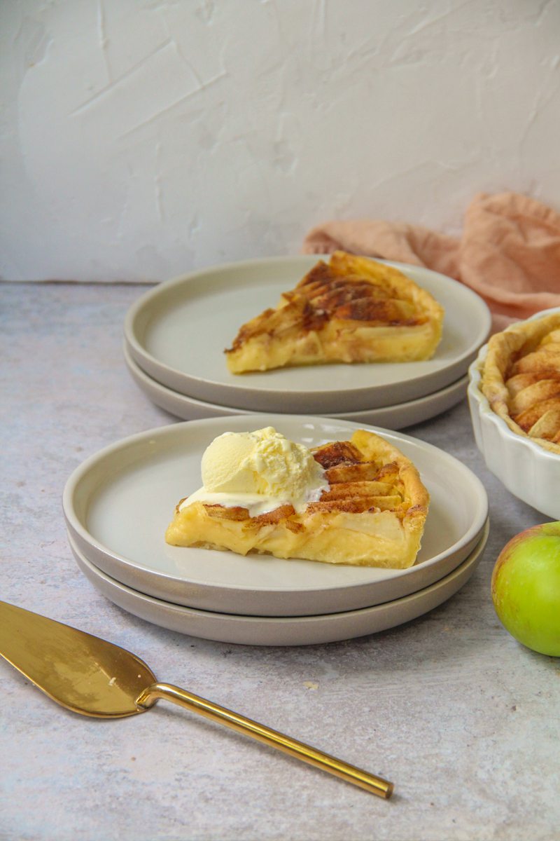 A slice of Portuguese Tarte de Maca sits on two stacked gray ceramic plates on a gray surface with a scoop of vanilla ice cream on top and a gold server and apple beside.