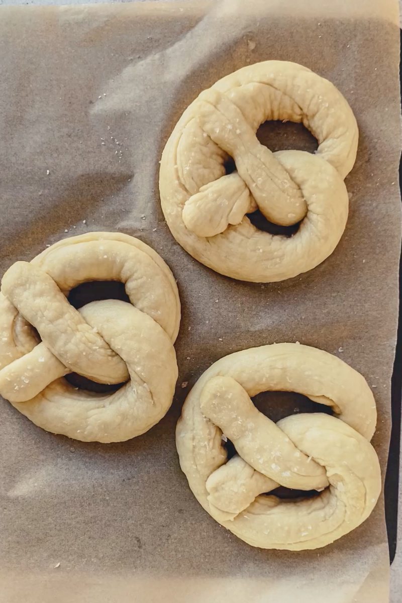 Unbaked pretzels sit on a lined baking tray topped with salt.