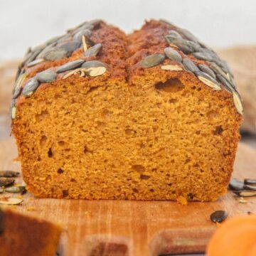 A close up of sliced pumpkin bread sits on a wooden board on a gray surface with small pumpkins beside.