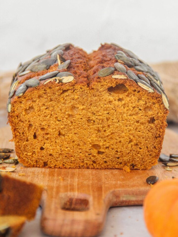 A close up of sliced pumpkin bread sits on a wooden board on a gray surface with small pumpkins beside.