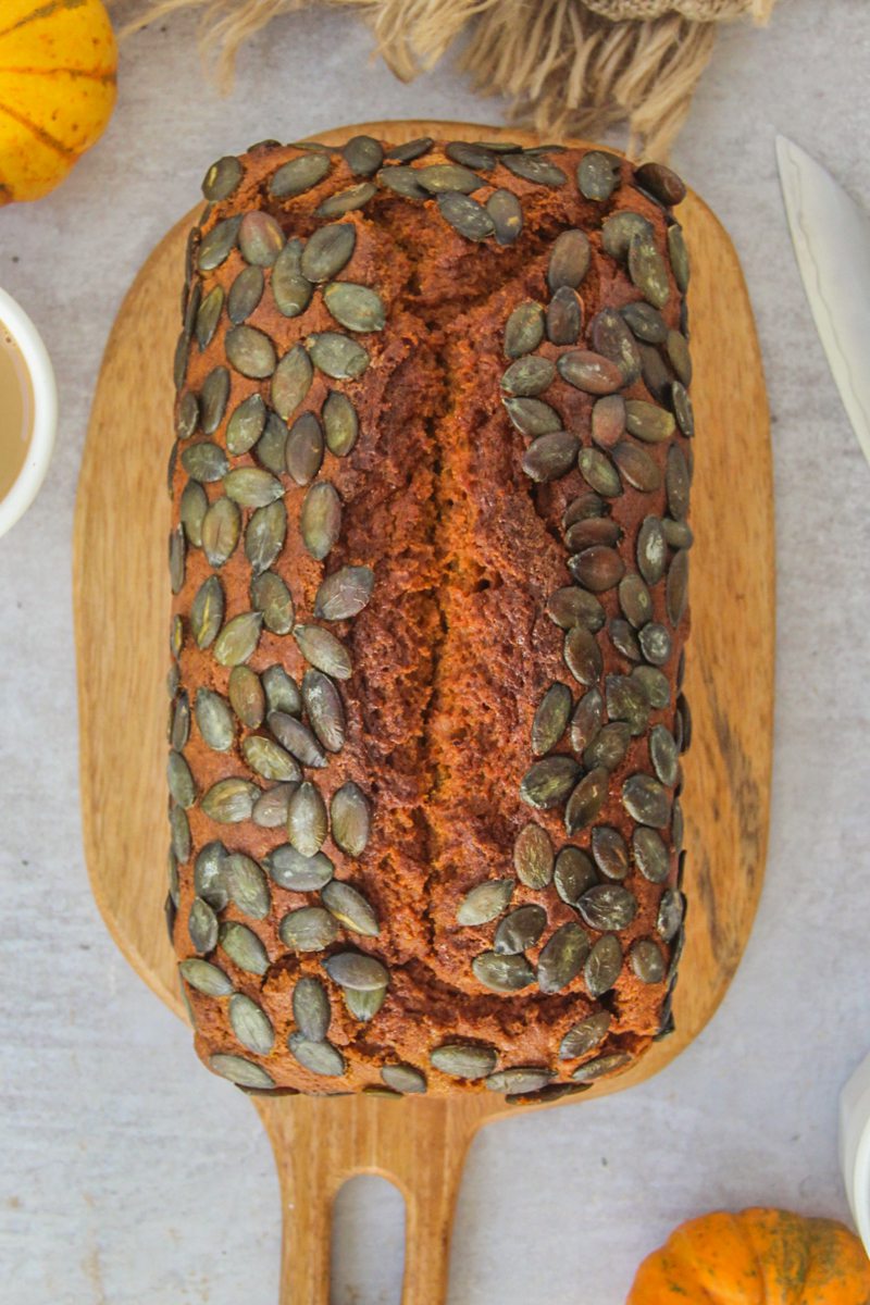 A loaf of pumpkin bread sits on a wooden board on a gray surface with small pumpkins beside.