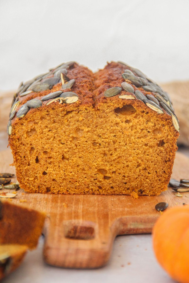 A close up of sliced pumpkin bread sits on a wooden board on a gray surface with small pumpkins beside.