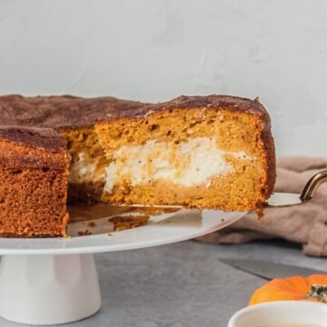 A slice of pumpkin cake with a cheesecake center is served up on a cake server whilst sitting on a white cake plate on a gray surface.