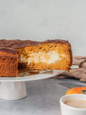 A slice of pumpkin cake with a cheesecake center is served up on a cake server whilst sitting on a white cake plate on a gray surface.