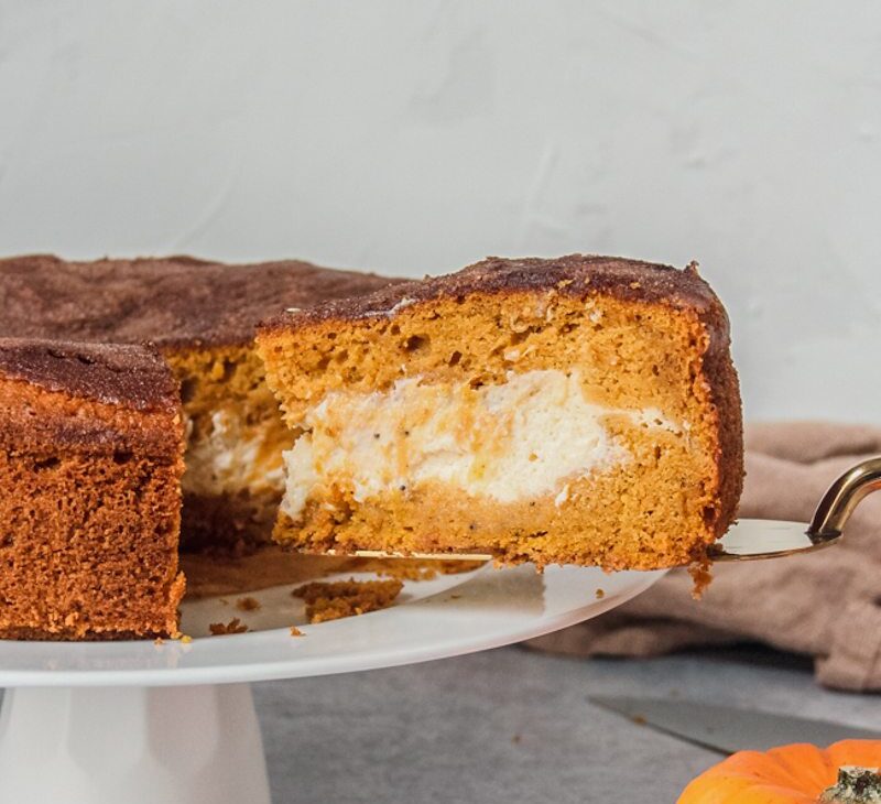 A slice of pumpkin cake with a cheesecake center is served up on a cake server whilst sitting on a white cake plate on a gray surface.