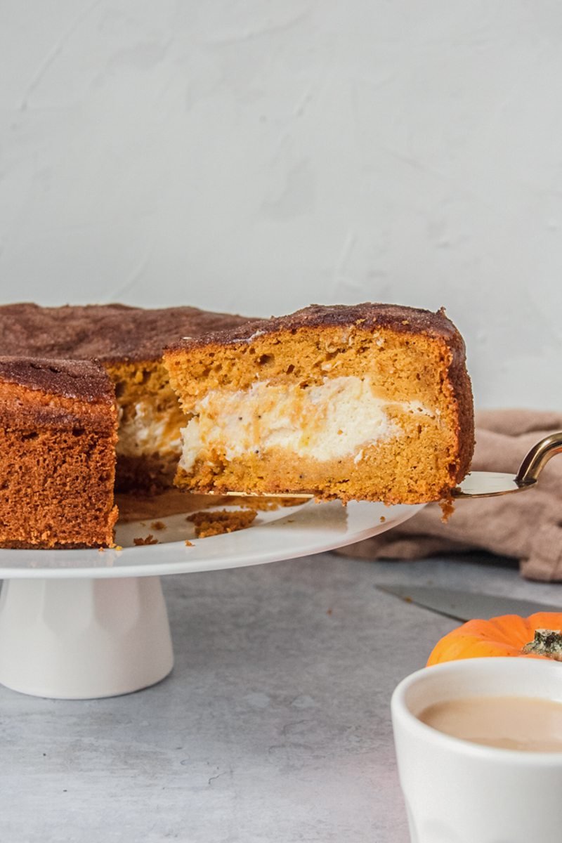 A slice of pumpkin cake with a cheesecake center is served up on a cake server whilst sitting on a white cake plate on a gray surface.