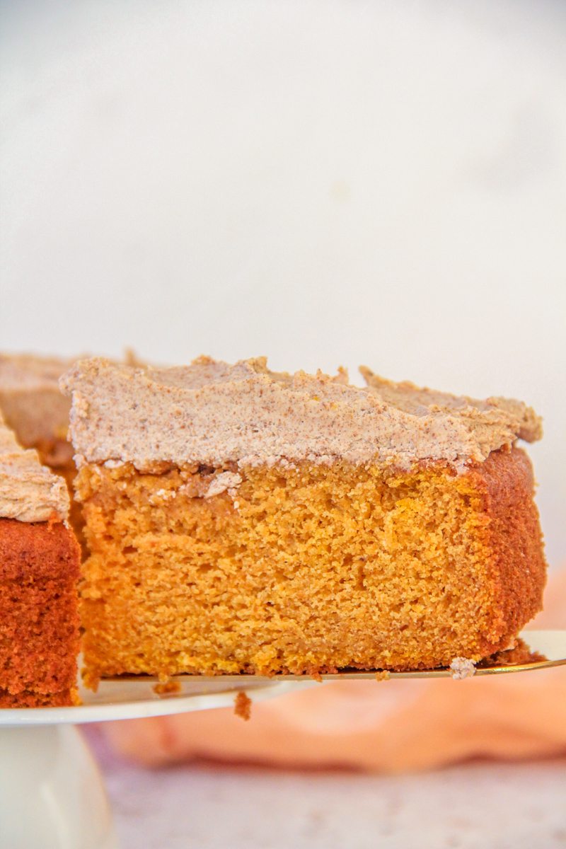 A slice of Pumpkin Cake with Coffee Buttercream sits on a cake server as the remaining cake sits on a cake stand.