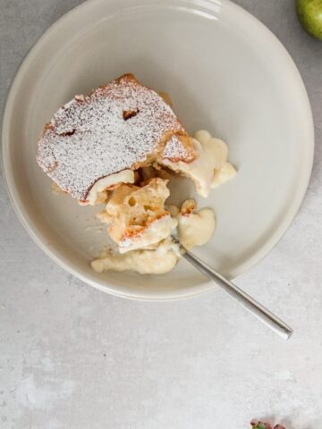 A spoonful of Pwdin Eva - Welsh Eve's Pudding sits on a rimmed ceramic plate with a small serving of custard on a gray surface.
