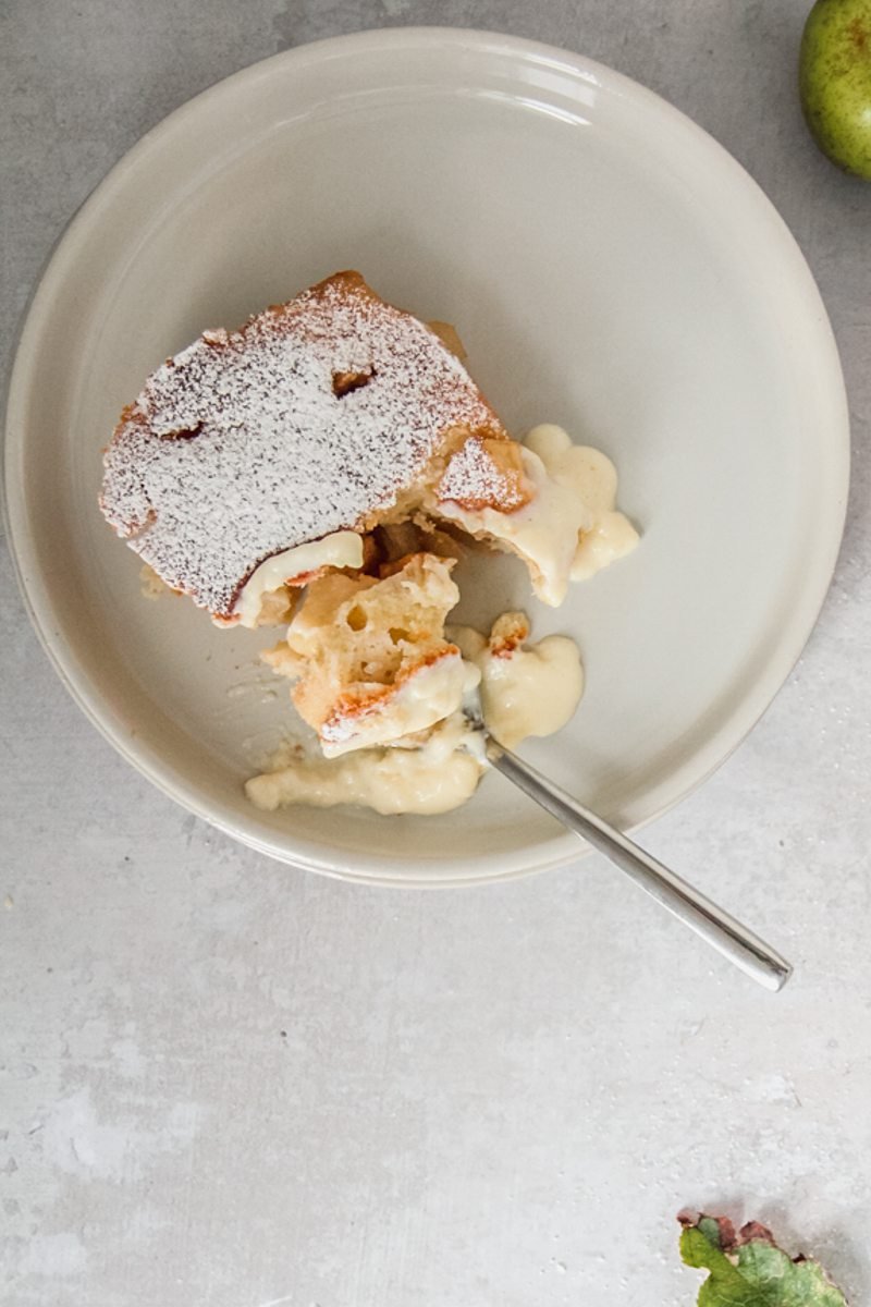 A spoonful of Pwdin Eva - Welsh Eve's Pudding sits on a rimmed ceramic plate with a small serving of custard on a gray surface.