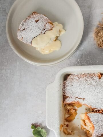 A spoonful of Pwdin Eva - Welsh Eve's Pudding sits on a rimmed ceramic plate with a small serving of custard with a portion of the remaining pudding sitting on a gray surface.