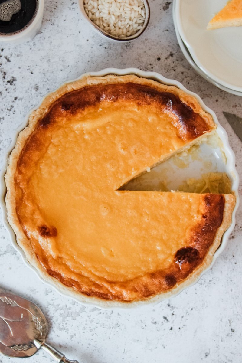 A Belgian Rijstaart or Rice Tart sits in it's white ceramic dish on a gray surface with one piece served up beside.