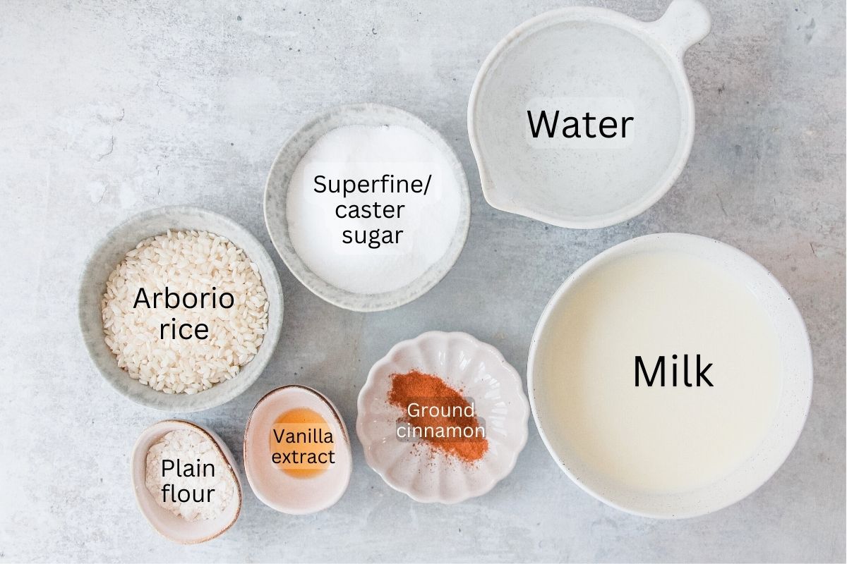 Ingredients for Rizogalo - Greek Rice Pudding sit in a series of bowls on a gray surface.