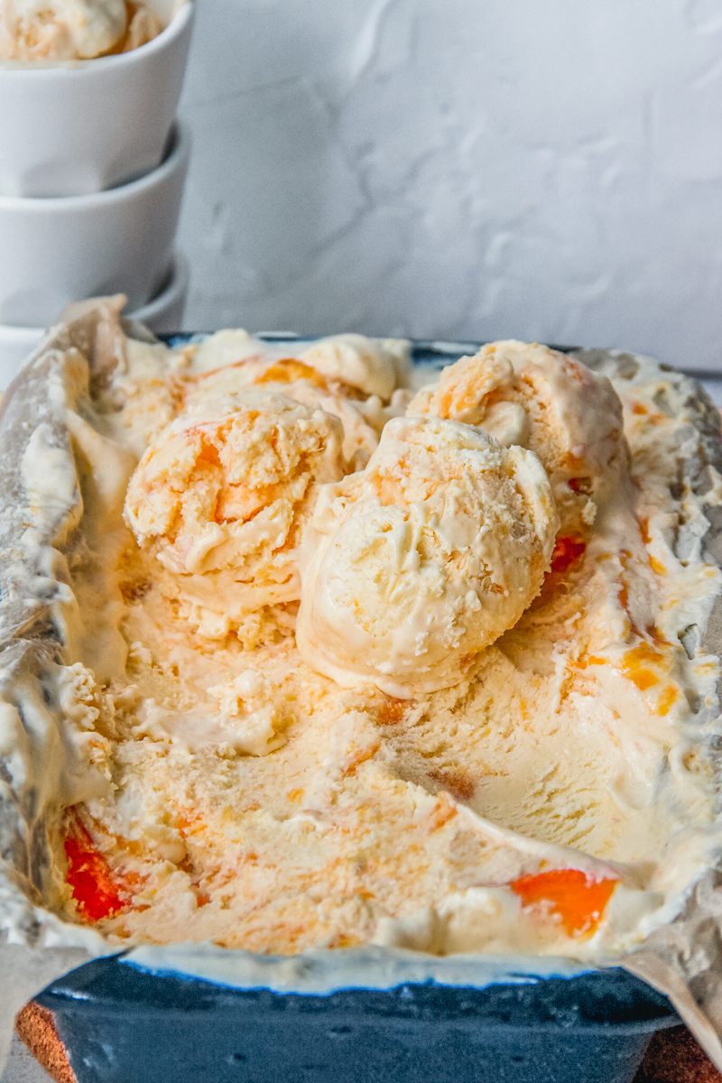 Scoops of roasted peach ice cream sit on top of the tin of ice cream in a lined metal loaf tin with cups stacked behind the tin.