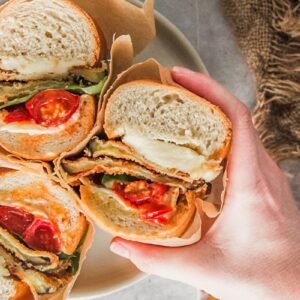 A roasted tomato and eggplant sandwich wrapped in parchment paper is picked up from a rimmed ceramic plate on a gray surface.