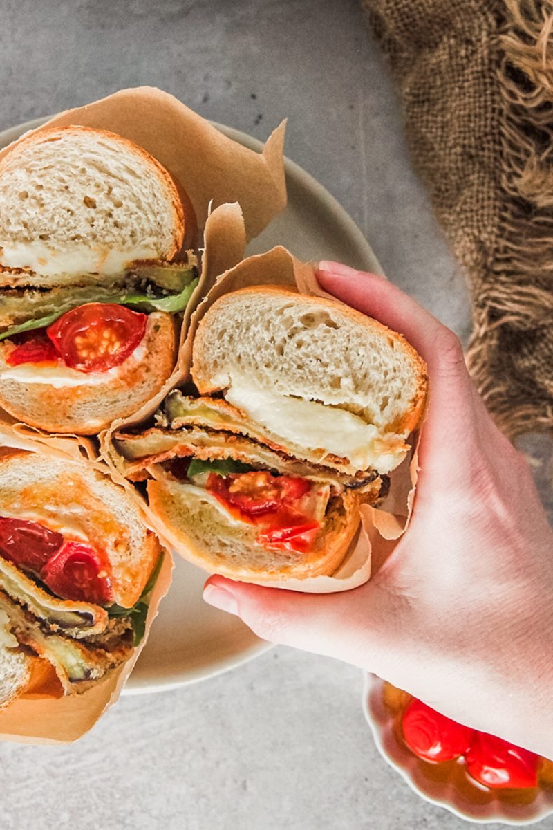 A roasted tomato and eggplant sandwich wrapped in parchment paper is picked up from a rimmed ceramic plate on a gray surface.