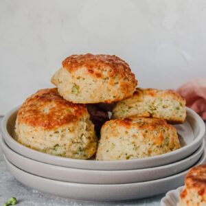 A stack of savory cheese biscuits sit on a stack of rimmed ceramic plate with butter on an individual plate on a gray surface.