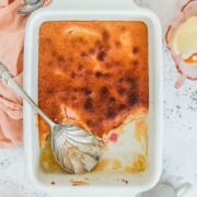A Rhubarb Sponge Pudding sits in a white ceramic baking dish on a light gray surface.