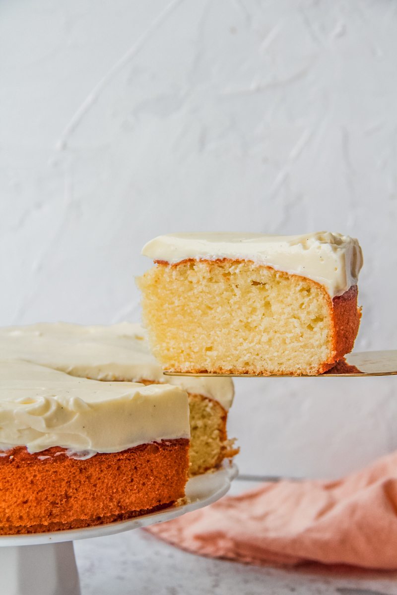 A slice of single layer vanilla cake sits on a cake server lifted from the remaining cake on a white ceramic stand on a light gray surface.