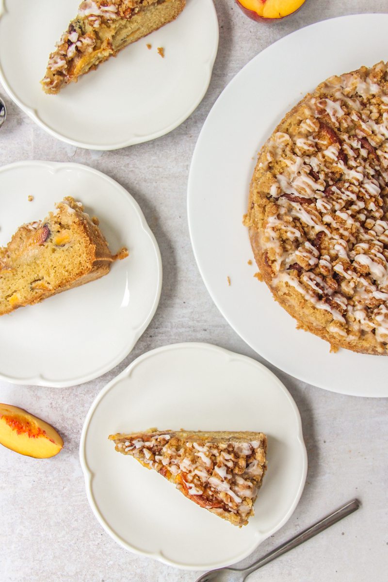 Slices of Peach Crumb Cake sit on round white ceramic plates on a gray surface with remaining cake beside.
