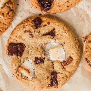 A close up of Smore cookies studded with marshmallow and chocolate chunks on parchment paper on a light gray background.