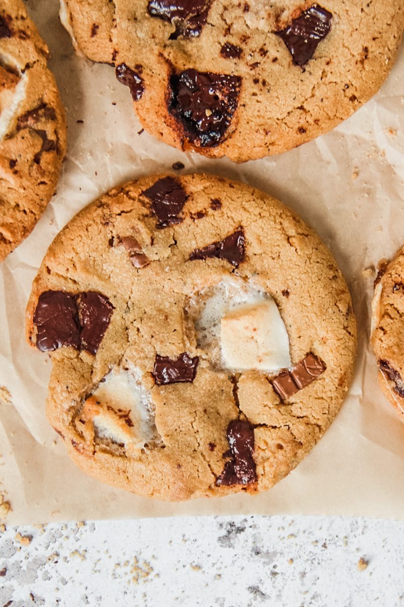 A close up of Smore cookies studded with marshmallow and chocolate chunks on parchment paper on a light gray background.