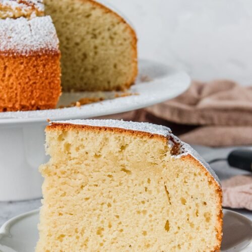 A slice of Spanish Olive Oil Cake sits on a white plate with the cake on a cake stand behind. A brown cloth beside it, all on a light grey background.