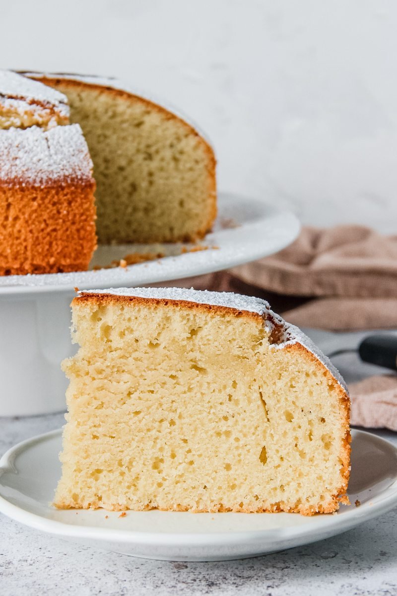 A slice of Spanish Olive Oil Cake sits on a white plate with the cake on a cake stand behind. A brown cloth beside it, all on a light grey background.