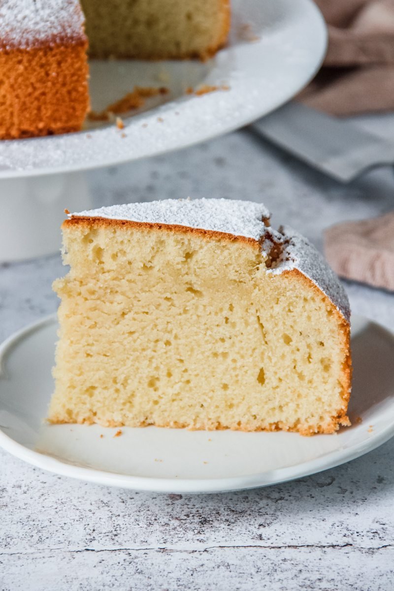 A slice of Spanish Olive Oil Cake sits on a white plate with the remaining cake sitting on a cake stand behind the slice on a light gray background.