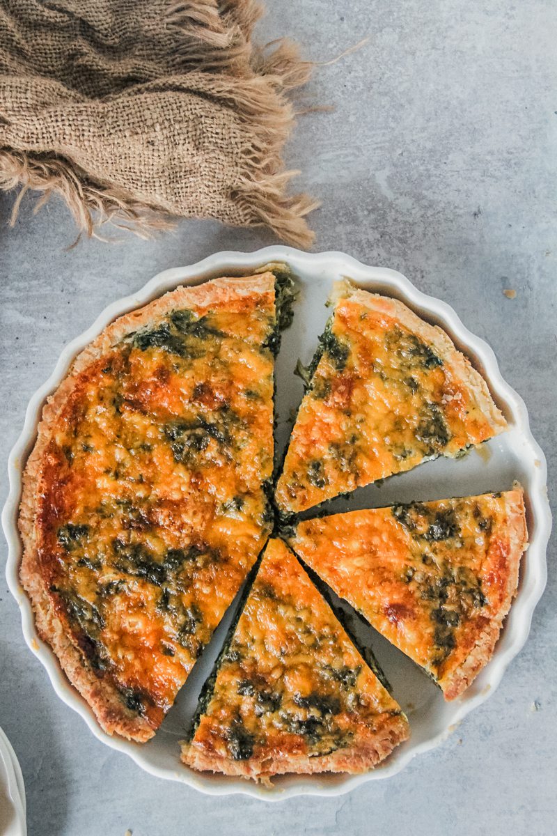 A spinach and cheese quiche sits in a white ceramic baking dish on a gray surface with a few slices cut out.