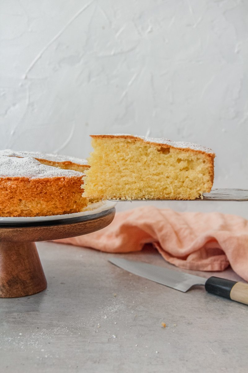 A slice of plain sponge cake sits on a cake server next to the remaining cake on a cake stand above a gray surface.