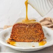 Toffee sauce is poured over Sticky Toffee Pudding on a serving plate on a gray surface.