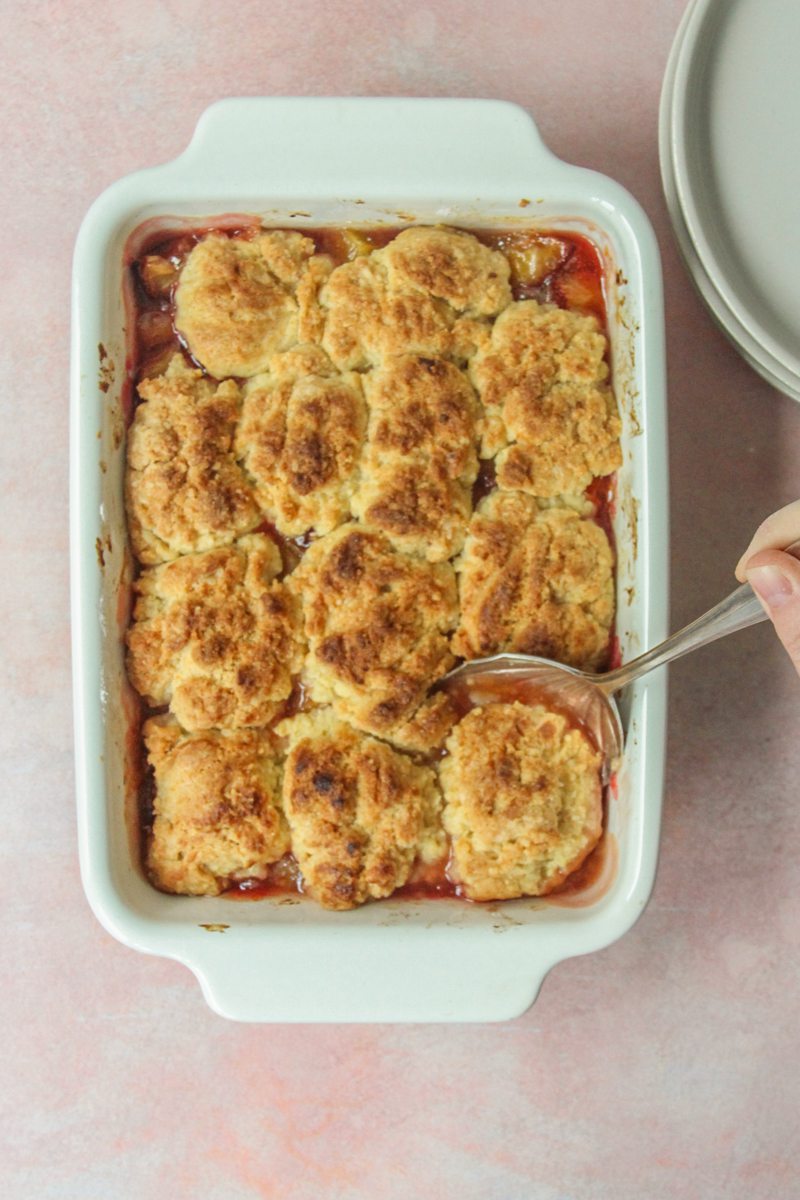 A stone fruit cobbler has a serving spoon dug into the corner of the dessert as it sits in a white ceramic baking dish on a light pink surface.