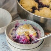 An individual serving of Canadian Blueberry Cobbler sits in a stack of blue ceramic plates on a light gray surface with the remaining cobbler sitting in the stainless steel saucepan behind.