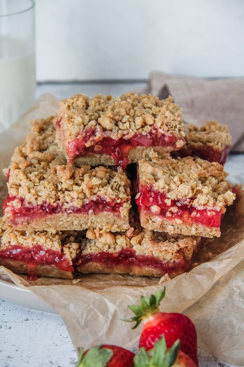 Strawberry Bars sit stacked on a plate with the juicy strawberry filling running down the oatmeal crumb base, sitting on a light grey background.