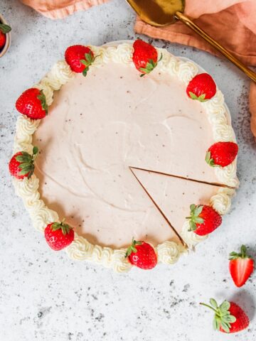 A no bake strawberry cheesecake topped with a whipped cream border and halved strawberries sits on a white plate on a light gray surface.