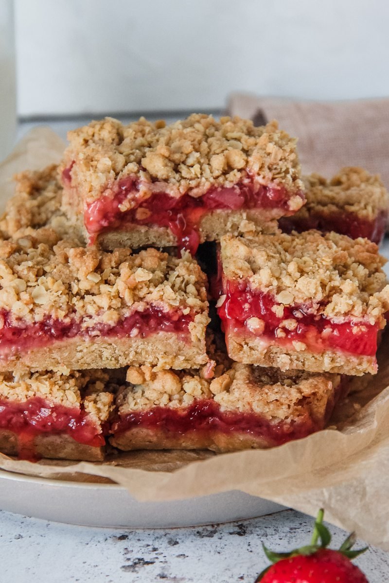 Strawberry Oat Bars sit stacked on a pkate with the juicy strawberry filling running down the oatmeal crumb base, sitting on a light grey background.