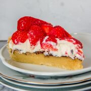 A slice of Danish strawberry tart sits on a stack of individual ceramic plates with the layers of tart visible on a gray surface.