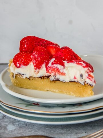 A slice of Danish strawberry tart sits on a stack of individual ceramic plates with the layers of tart visible on a gray surface.